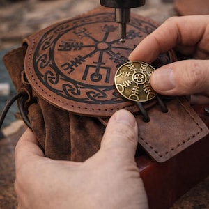 Peut inclure: Une pochette en cuir marron avec un motif circulaire et un bouton doré en cours d'attache. La pochette présente un motif en relief détaillé. Le bouton a un symbole unique. L'image montre un gros plan du processus de fabrication.
