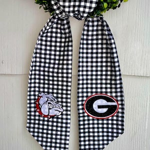 May include: A decorative wreath with a black and white checkered bow and streamers. The streamers feature the University of Georgia Bulldogs logo and mascot. The wreath is adorned with green foliage.