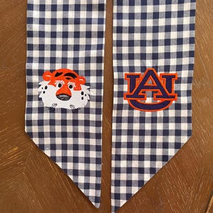 May include: A pair of blue and white gingham fabric graduation sashes with embroidered logos. One sash features a cartoon tiger face and the other features the letters "AU" in orange and blue.