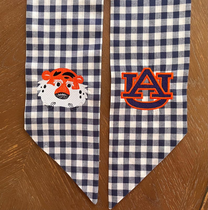 May include: A pair of blue and white gingham fabric graduation sashes with embroidered logos. One sash features a cartoon tiger face and the other features the letters "AU" in orange and blue.