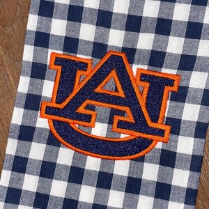 May include: A white and navy blue gingham kitchen towel with an embroidered orange and navy blue "AU" logo.