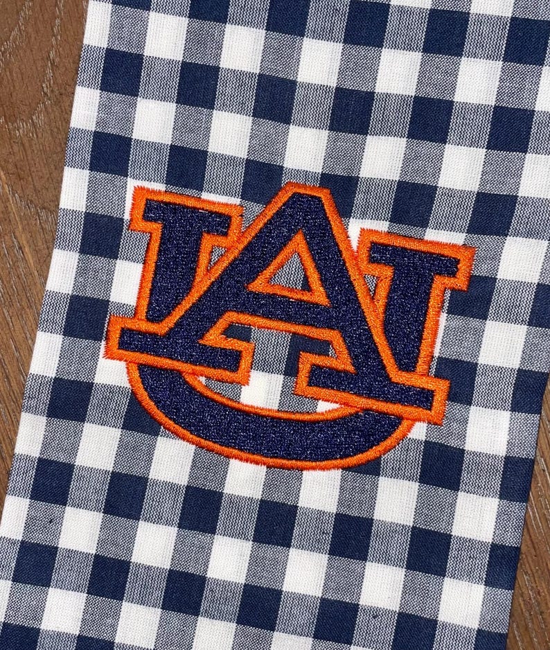 May include: A white and navy blue gingham kitchen towel with an embroidered orange and navy blue "AU" logo.