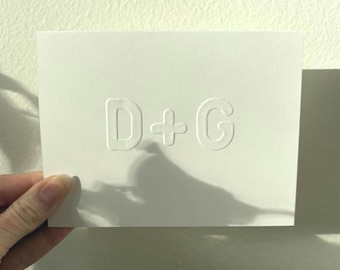 May include: A rectangular white card featuring the embossed letters D and G separated by a plus sign in the centre. The design is created through a debossed technique, resulting in a subtle three-dimensional appearance on the smooth paper surface.