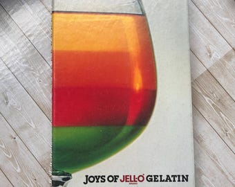 Op de afbeelding: Een spiraalgebonden kookboek getiteld "Joys of Jell-O Gelatin" toont een kleurrijk gelaagd gelatinedessert in een groot glas. Het dessert heeft groene, rode, oranje en gele lagen. Het boek ligt op een licht oppervlak met houtnerf.