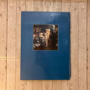 Op de afbeelding: Een blauw hardcover boek met een rechthoekige afbeelding. De afbeelding toont een sc&egrave;ne met verschillende mensen en een robot. De achtergrond is een houten oppervlak.
