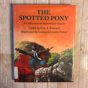 May include: A hardcover book titled "THE SPOTTED PONY" with an orange cover. The book is a collection of Hanukkah stories retold by Eric A. Kimmel and illustrated by Leonard Everett Fisher. The cover illustration depicts a man in a carriage pulled by a horse.