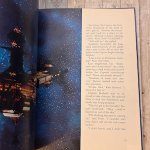 Op de afbeelding: Een open boekpagina met een sciencefiction-illustratie van een ruimteschip en een ruimtestation tegen een sterrenachtergrond. Tekst op de rechterpagina beschrijft de sc&egrave;ne, met het nummer 19 onderaan.