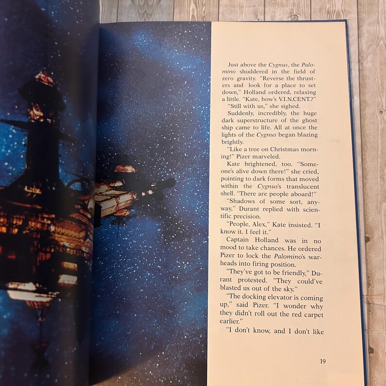 Op de afbeelding: Een open boekpagina met een sciencefiction-illustratie van een ruimteschip en een ruimtestation tegen een sterrenachtergrond. Tekst op de rechterpagina beschrijft de sc&egrave;ne, met het nummer 19 onderaan.