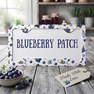 Puede incluir: Un marco de matrícula rectangular con un patrón repetitivo de arándanos azules y hojas verdes sobre un fondo blanco. El texto BLUEBERRY PATCH está impreso en letras mayúsculas de color azul oscuro en el centro del marco.