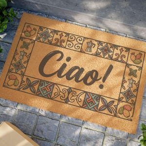 May include: A rectangular tan coir doormat featuring the word Ciao! in dark brown script in the center. The border is decorated with a repeating pattern of espresso makers, prickly pear fruits, and geometric shapes in muted green and red tones.