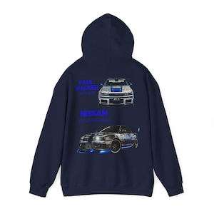 Puede incluir: Sudadera con capucha azul marino con un Nissan Skyline GT-R blanco impreso en la parte posterior. El coche está rodeado de un brillo azul. El texto "Paul Walker" y "Nissan Skyline GT-R" está impreso en azul arriba y abajo del coche.