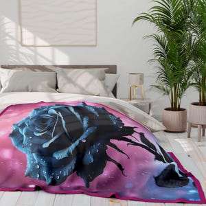 Könnte beinhalten: Eine weiche Decke mit einer großen, dunkelblauen Rose mit Wassertropfen, vor einem rosa-violetten Farbverlauf. Die Decke hat einen magentafarbenen Rand und ist über einem Bett drapiert.