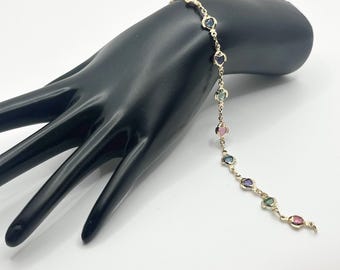 May include: A gold-toned bracelet featuring a series of colorful, heart-shaped gemstones. The bracelet is displayed on a black hand, showcasing the delicate design and vibrant hues of the stones. The gemstones include red, blue, purple, green, and pink.