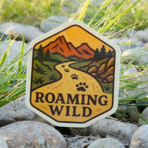 May include: A hexagonal sticker with a nature scene. The design features a winding path with paw prints, leading to mountains under an orange sky. The text "ROAMING WILD" is at the bottom. The sticker is on a bed of rocks and grass.