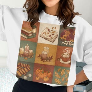 Puede incluir: Sudadera blanca con un diseño de patchwork con ilustraciones de temática otoñal. El diseño incluye libros, velas, calabazas, flores y una taza de café. El tema general es el otoño y el ambiente acogedor.