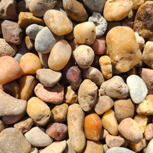 Puede incluir: Primer plano de una variedad de piedras de río lisas y redondeadas en tonos marrones, grises y rojos. Las piedras están dispuestas en un patrón aleatorio, creando un aspecto natural y texturizado.