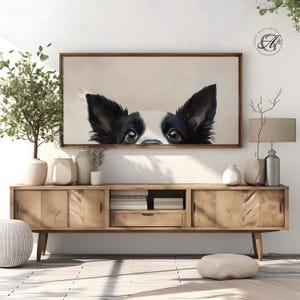 May include: A black and white border collie dog peeks over the top of a framed canvas print. The dog's eyes are visible, and the print is hanging on a white wall above a wooden entertainment center.