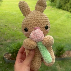 Puede incluir: Un peluche de ganchillo color canela con forma de conejo, orejas largas, ojos de plástico negro y una pequeña nariz bordada en rosa. El conejo sostiene una pequeña flor rosa texturizada con un tallo verde claro de lana suave.