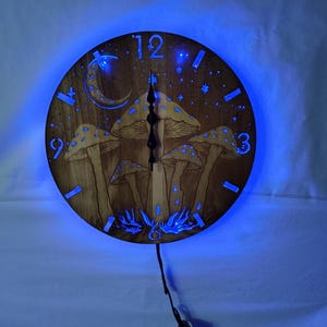 Può includere: Un orologio in legno con un design a forma di fungo e luna. L'orologio ha una luce LED blu che illumina il design. L'orologio ha un diametro di 30 cm.