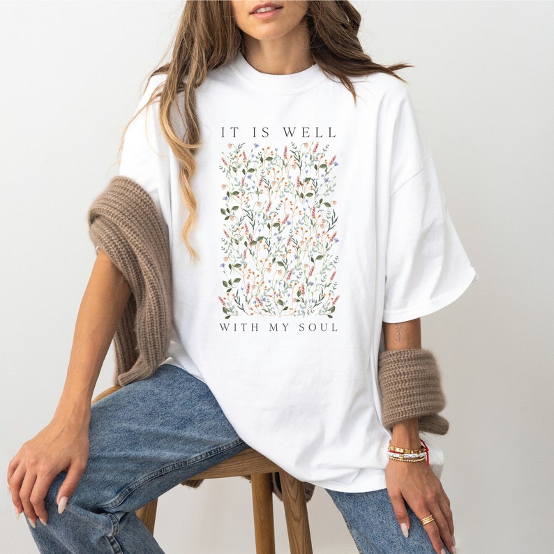 Op de afbeelding: Wit oversized T-shirt met een bloemenprint en de tekst "It is well with my soul".
