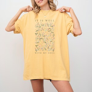 Op de afbeelding: Een oversized geel t-shirt met een bloemenprint en de tekst "It is well with my soul".