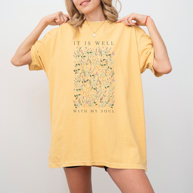 Op de afbeelding: Een oversized geel t-shirt met een bloemenprint en de tekst "It is well with my soul".
