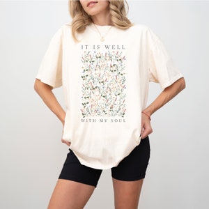 Op de afbeelding: Een cr&egrave;mekleurig T-shirt met een bloemenprint en de tekst "It is well with my soul".