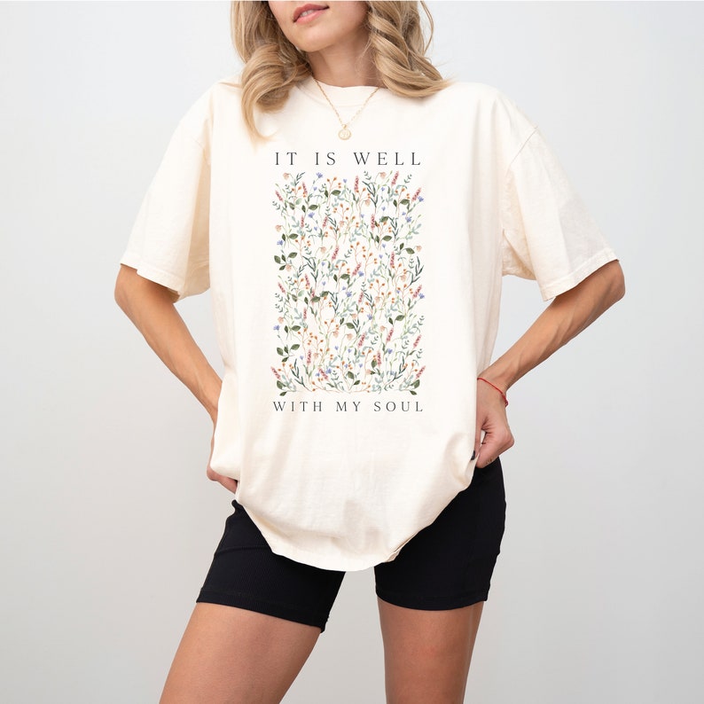 Op de afbeelding: Een cr&egrave;mekleurig T-shirt met een bloemenprint en de tekst "It is well with my soul".