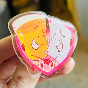 May include: A heart-shaped, glittery acrylic pin featuring cartoon characters. One character is a smiling glass of orange juice, the other is a smiling stack of paper. The pin has a pink border and a clear, glittery background.