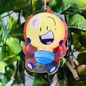 May include: A clear acrylic charm featuring a cartoon lightbulb character with a yellow body, orange outline, and blue diaper. The charm has a gold-colored keyring and is set against a backdrop of green foliage.