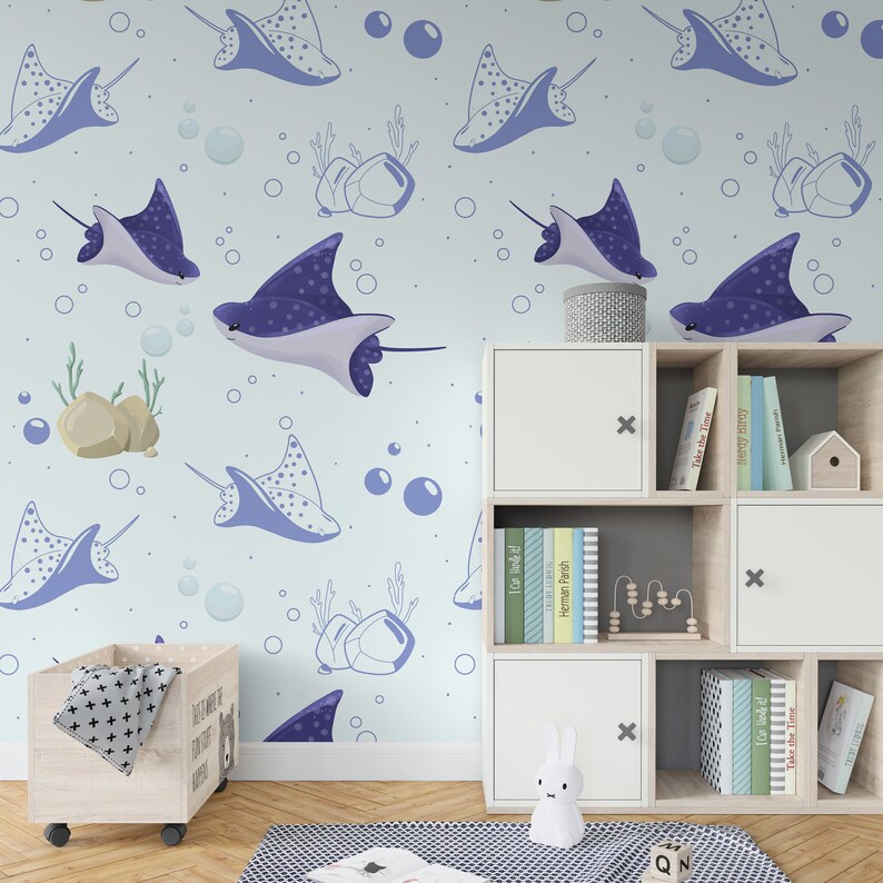 K&ouml;nnte beinhalten: Eine hellblaue Kinderzimmertapete mit einem sich wiederholenden Muster aus blauen Rochen, Blasen und Korallen. Die Tapete ist an einer Wand mit einem wei&szlig;en B&uuml;cherregal und einer Holzkiste f&uuml;r Spielzeug angebracht.