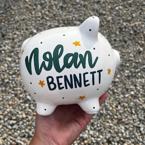May include: White ceramic piggy bank with the name "Nolan Bennett" in green script and navy block letters. The bank is decorated with yellow star accents and small green dots.