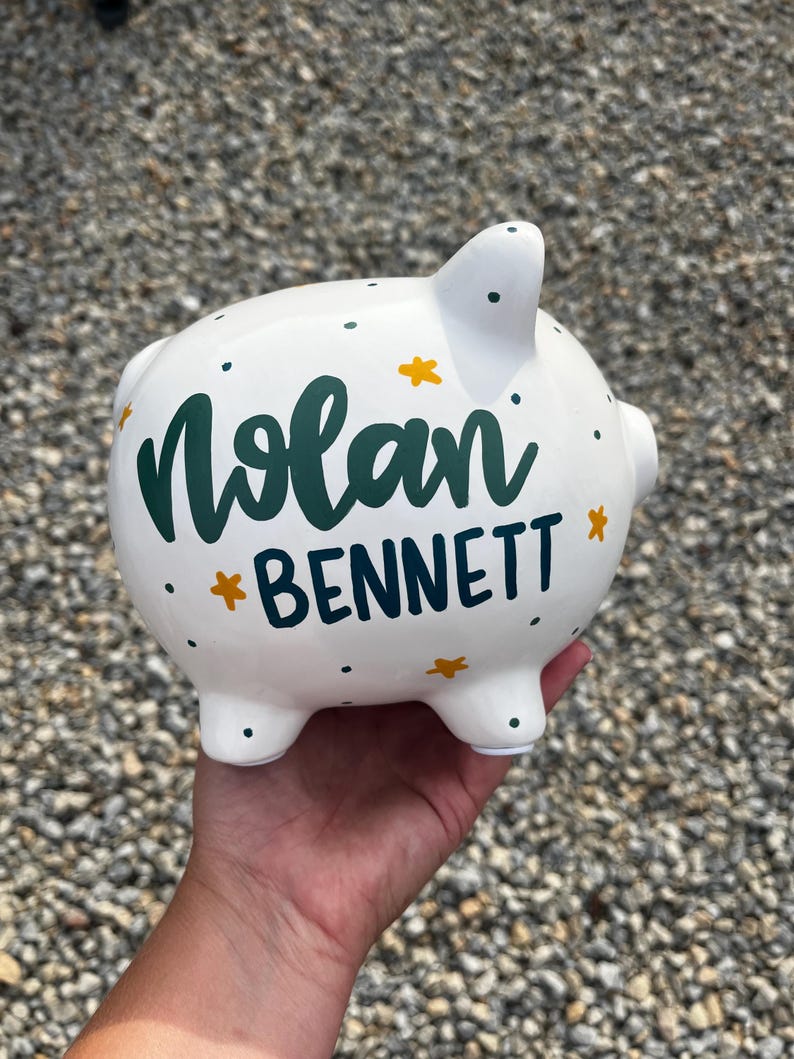May include: White ceramic piggy bank with the name "Nolan Bennett" in green script and navy block letters. The bank is decorated with yellow star accents and small green dots.