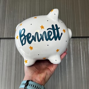 May include: White ceramic piggy bank decorated with teal lettering that spells "Bennett", surrounded by yellow stars and gray dots. The piggy bank has four small feet and a slot on top for inserting coins.