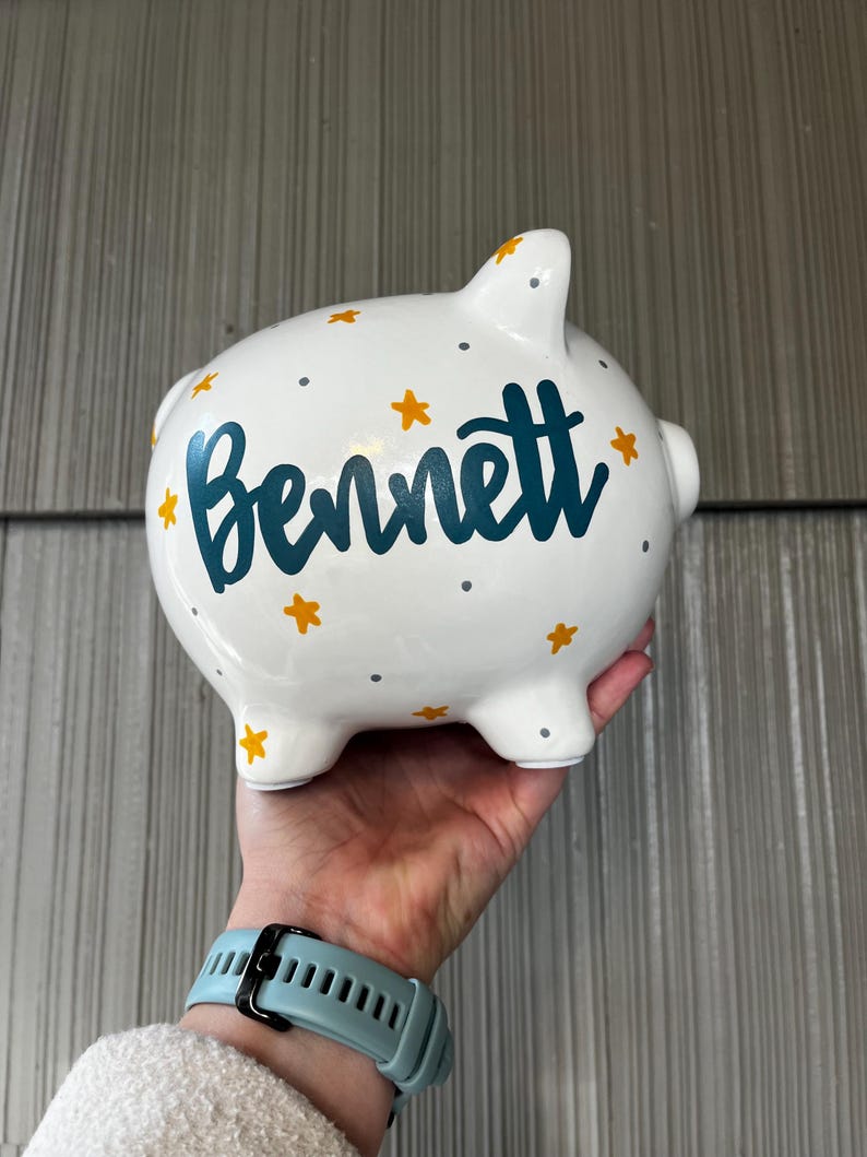 May include: White ceramic piggy bank decorated with teal lettering that spells "Bennett", surrounded by yellow stars and gray dots. The piggy bank has four small feet and a slot on top for inserting coins.