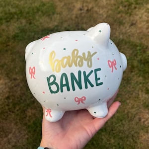 May include: White ceramic piggy bank with the words "baby BANKE" in gold and green lettering. The bank is decorated with pink bows and colorful polka dots. A charming and decorative item for saving.