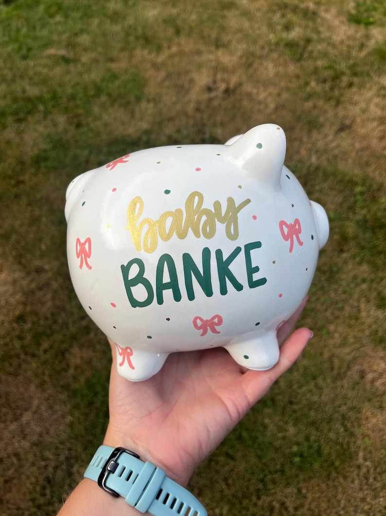 May include: White ceramic piggy bank with the words "baby BANKE" in gold and green lettering. The bank is decorated with pink bows and colorful polka dots. A charming and decorative item for saving.