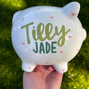 May include: White ceramic piggy bank with the name "Tilly Jade" in green and teal lettering. The bank is decorated with small pink hearts. The piggy bank has four feet, a tail, and two ears.
