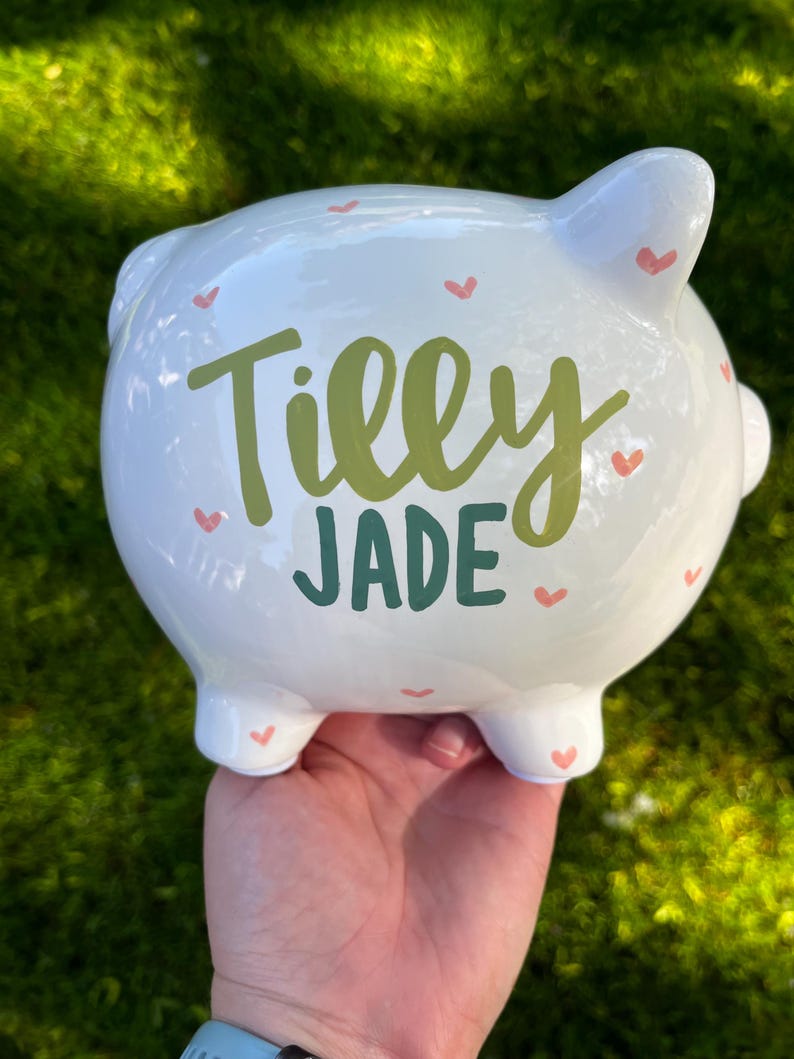 May include: White ceramic piggy bank with the name "Tilly Jade" in green and teal lettering. The bank is decorated with small pink hearts. The piggy bank has four feet, a tail, and two ears.