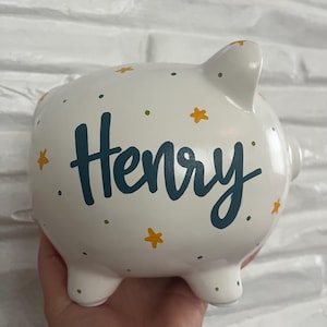 May include: White ceramic piggy bank with the name "Henry" in teal script. The bank is decorated with yellow star and green dot accents. The piggy bank has four small feet and a slot on top for inserting coins.