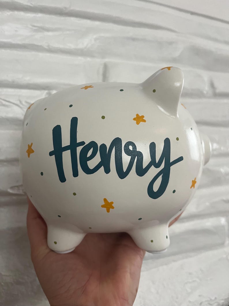 May include: White ceramic piggy bank with the name "Henry" in teal script. The bank is decorated with yellow star and green dot accents. The piggy bank has four small feet and a slot on top for inserting coins.