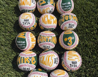 Può includere: Una collezione di quattordici palle da baseball bianche con cuciture rosse, ognuna caratterizzata da pannelli verdi dipinti a mano e scritte gialle. Le palle sono personalizzate con vari nomi, numeri, l'anno 2026 e la frase Hawk Eyes Full Hearts Can't Lose.