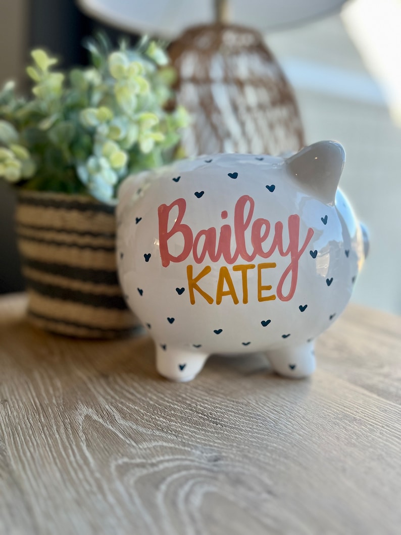 May include: White ceramic piggy bank with the name "Bailey KATE" in pink and gold lettering. The bank is decorated with small navy blue hearts. The piggy bank has four small feet and a slot on top.