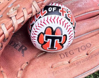 May include: A tan leather baseball glove with visible lacing and the text DFG 700 premium grade leather, holding a white baseball with red stitching. The ball features a black and orange paw print logo with a letter T and the text Hall of Fame.