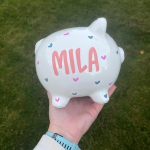 May include: White ceramic piggy bank with the name "MILA" in pink lettering. The bank is decorated with pink and teal hearts. The piggy bank is held in a hand against a green background.