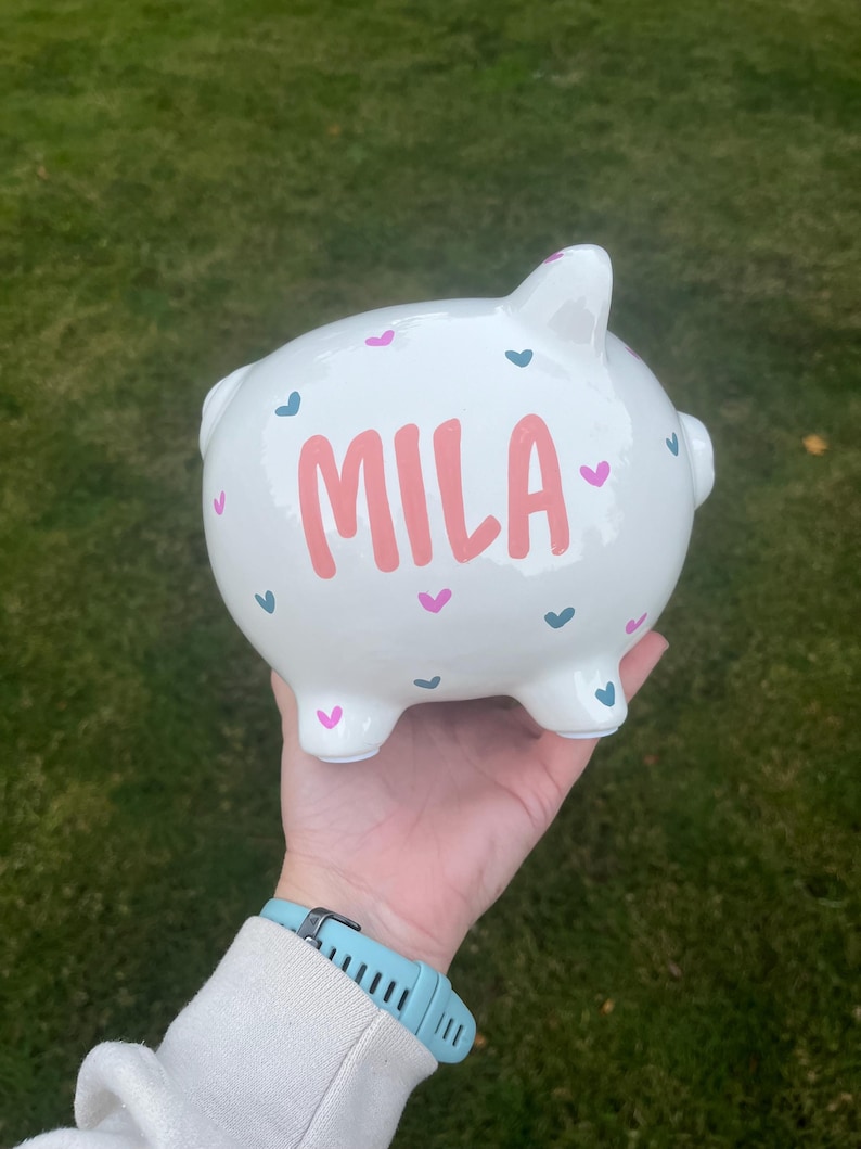 May include: White ceramic piggy bank with the name "MILA" in pink lettering. The bank is decorated with pink and teal hearts. The piggy bank is held in a hand against a green background.