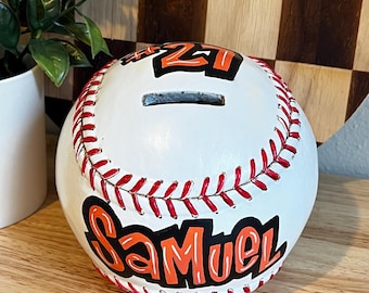 Puede incluir: Una hucha de cerámica con forma de pelota de béisbol blanca con costuras rojas y el nombre "Samuel" en letras naranjas y negras. El número "21" también se muestra en naranja y negro. Una ranura está cortada para monedas.
