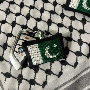 May include: A green, white, and black embroidered keychain with a crescent moon and star design. The keychain is on a white and black patterned fabric.