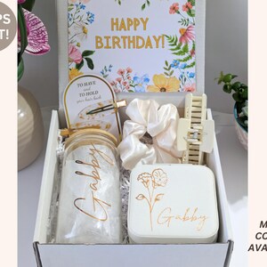 May include: A gift box with a floral "Happy Birthday" card, a jar with "Gabby" in gold script, a white jewelry box with a floral design, a hair claw clip, and a scrunchie. The box also contains a hair clip and a small card.