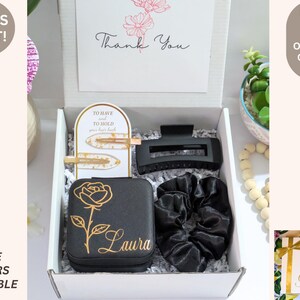 May include: A gift box containing a black jewelry case with a gold rose design and the name "Laura", a black hair clip, two gold hair clips, and a black scrunchie. A card reads "Thank You".